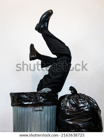 Businessman in Suit Stuck Upside Down in Metal Trash Can Next to Garbage Bag Pile. Concept of Over a Barrel. Thrown Away by Capitalism and Greed. Royalty-Free Stock Photo #2163638725
