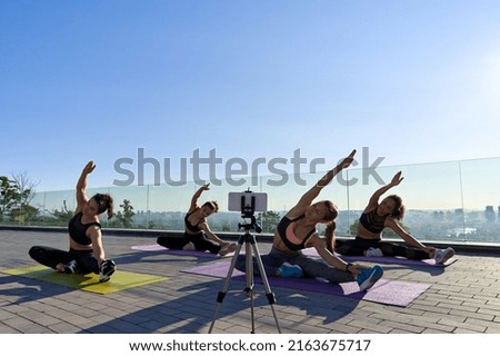 Diverse fit sporty young women doing yoga exercise stretching social media online stream class shooting group training video blog on mobile phone camera. Remote fitness tutorial vlog workout outdoor.