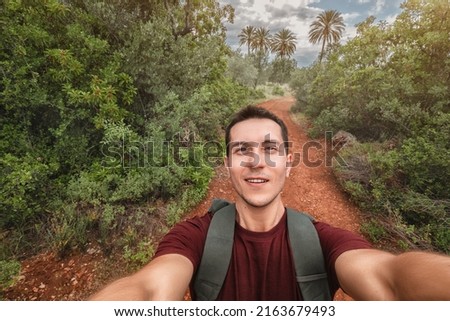 Young happy man takes selfie photo during his awesome hiking trek somewhere in Africa national park, walking by red soil footpath in jungle