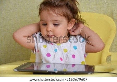 a little cute girl with blue eyes, a slider sits in a child seat and plays a tablet, watches cartoons. early childhood. Beautiful child little girl with ponytails playing with a tablet 