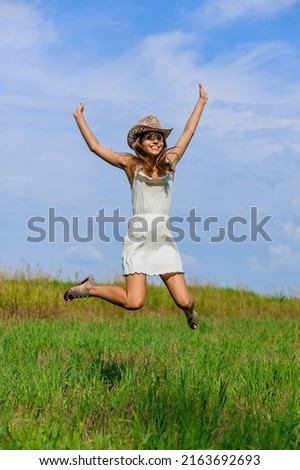 A beautiful laughing young woman in a white dress and a cowboy hat jumping against the green grass on a summer sunny day.
