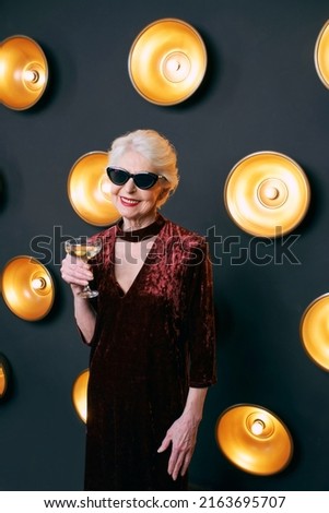 senior stylish elegant woman in sunglasses with glass of sparkling wine on lamps dark background. Party, fashion, celebration, anti age concept 