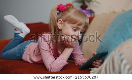 Little one school kid girl children using mobile phone technology device lying on couch alone at home. Child toddler hold smartphone watching funny cartoons, chatting, play online game. Tech addiction