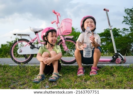 Happy cute little girl and her sister sit on the roadside lawn and drink water from a bottle near a bike in the park. Kids resting after biking. Healthy Summer Activities for Kids.