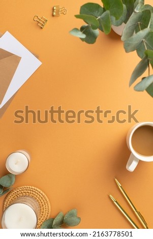 Top view vertical photo of workplace cup of coffee paper sheet envelops pens binder clips candles rattan serving mat and vase with eucalyptus on isolated orange background with copyspace in the middle