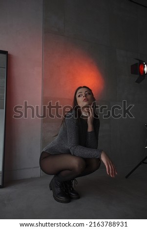 young teen girl in a shiny silver bodysuit is sitting fashion in profile on floor with hand near face on the gray wall background in loft interior with red studio light. fashion concept, free space