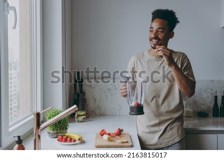 Young smiling african american man 20s in casual clothes prepare fruit cocktail eat strawberry using mixer blender cooking food in light indoor kitchen at home alone. Healthy diet lifestyle concept. Royalty-Free Stock Photo #2163815017