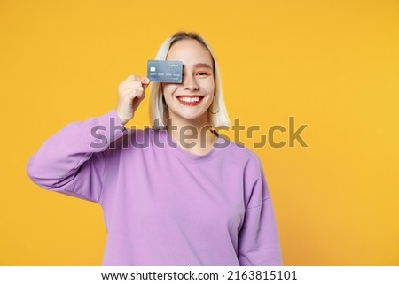 Smiling young blonde caucasian happy woman bob haircut bright makeup wearing casual basic purple shirt holding in hand covering eye with credit bank card isolated on yellow background studio portrait Royalty-Free Stock Photo #2163815101