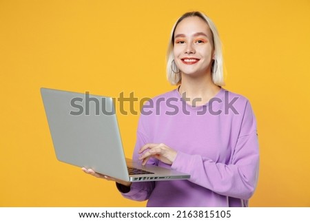 Smiling young blonde caucasian woman 20s with bob haircut bright makeup wearing basic purple shirt using modern laptop pc computer typing keyboard isolated on yellow color background studio portrait