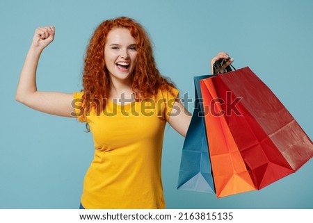 Young surprised happy redhead woman 20s in yellow t-shirt holding package bags with purchases after shopping do winner gesture isolated on plain light pastel blue background. People lifestyle concept