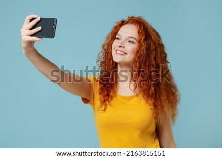 Young smiling cheeful happy redhead woman 20s wearing yellow t-shirt doing selfie shot on mobile cell phone post photo on social network isolated on plain light pastel blue background studio portrait.