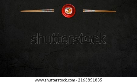 Kitchen table with Japanese sushi roll on round red plate with wooden chopsticks. Presented on the black concrete background, with black bottom center copy space. Table top view.