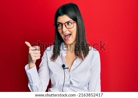 Young hispanic woman using lavalier microphone pointing thumb up to the side smiling happy with open mouth 