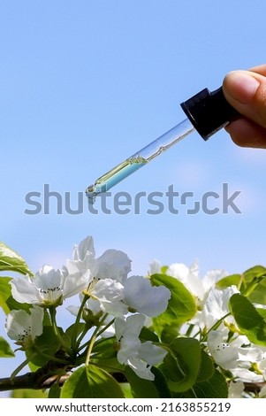 Closeup of the hand with the dropper with a drop of skincare serum over the beautiful white flowers against the blue sky