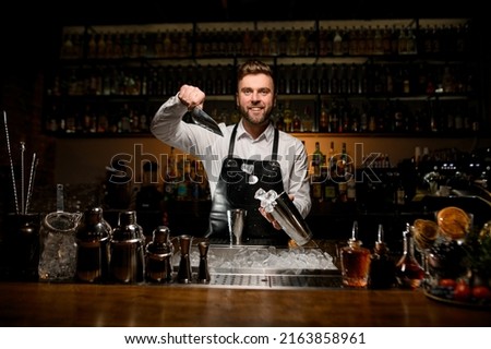 great view of smiling man bartender skillfully pouring ice cubes from metal scoop into shaker cup. Different bar equipment on the bar counter at foreground Royalty-Free Stock Photo #2163858961
