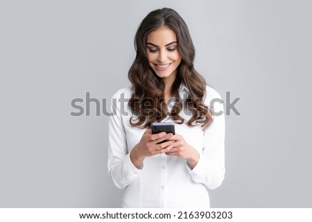 Young woman reading message on mobile phone and smiling. Happy businesswoman with smartphone, standing on grey background. Royalty-Free Stock Photo #2163903203
