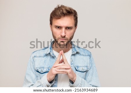 Young hipster male with stubble in a white t-shirt and light blue denim shirt. Emotion - desperately tired, angry, expects, listens displeasedly, hands folded in a castle. On a light gray background