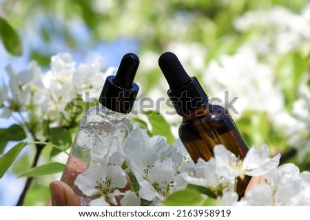 Closeup of the transparent and brown skincare glass bottle containers with dropper among the beautiful white flowers in springtime