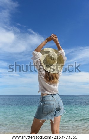 A woman is resting on the beach. She is wearing a straw hat, denim shorts and a white shirt. Travel and good rest, happy holidays, tourism, Philippines, summer season. Rear view, vertical