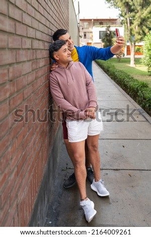 Vertical photo of a homosexual couple standing between a building and a park taking a selfie