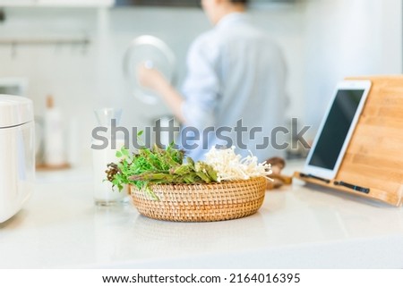 Close-up of a basket of fresh vegetables on family kitchen counter, with a housewife cooking in the background - stock photo