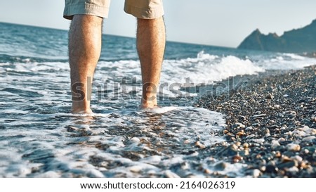 Man wearing shorts, walking barefoot along the seashore. Male legs walks on pebble beach along the shore near the water with waves, low section. Wellness, freedom and travel in summertime concept. Royalty-Free Stock Photo #2164026319