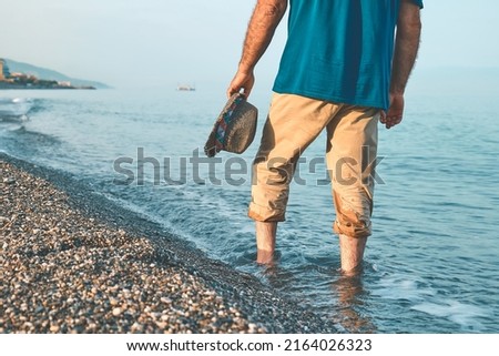 Man wearing shorts, walking barefoot along the seashore. Male legs walks on pebble beach along the shore near the water with waves, low section. Wellness, freedom and travel in summertime concept. Royalty-Free Stock Photo #2164026323