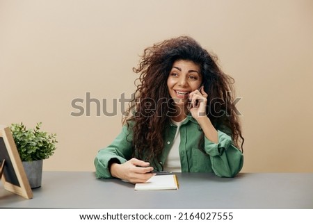 Happy smiling tanned curly Latin lady talking with friend in call with phone sit at the table isolated over pastel beige background look aside. Copy space Mockup Banner. Concept cell communication