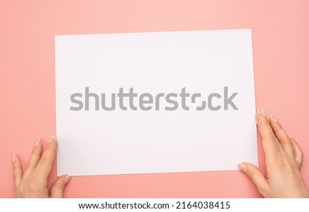 Mockup paper. Overhead shot of woman hands with blank paper sheet on pink table. Human hands holding mockup white advertising card. Isolated on pink background
