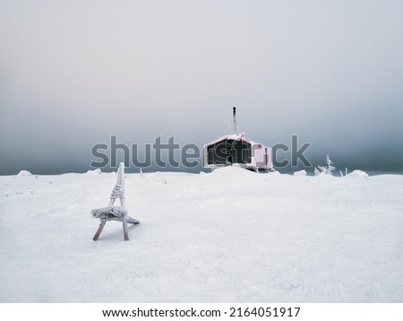 Selective focus. Wooden bench against the background of a lonely red guest house on a snow-covered slope in winter under dramatic sky. Double house on the mountain Volodyanaya Kandalaksha in Russia. 