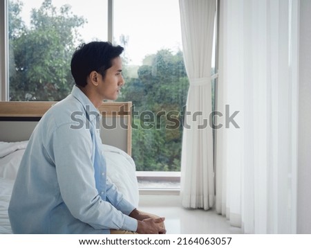 Thoughtful Asian man in denim shirt sitting on the bed feeling lonely, looking away with sadness and thinking in the bedroom at home near the glass window in the morning. Royalty-Free Stock Photo #2164063057