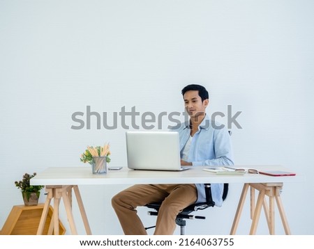 Handsome young Asian man in denim shirt using laptop on table on white clean space wall background in home office. Learning, working, startup with small business, stock, financial, investment concept.
