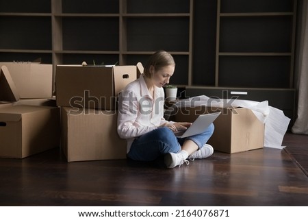 Woman sits on floor near cardboard boxes use laptop make order via website for transport services on move-out day, buy furniture on internet, search repairs company or interior design ideas concept