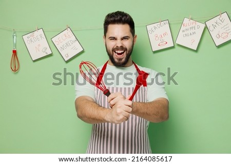 Young expressive happy cheerful male chef confectioner baker man 20s in striped apron hold whisk silicone egg brush isolated on plain pastel light green background studio portrait Cooking food concept
