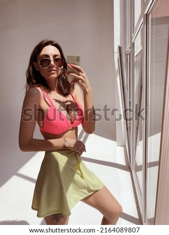 Fit tanned woman in light green beach skirt, sunglasses, bright pink swimsuit bra, summer fashion stylish wear, take photo selfie on phone in mirror for social media, stories, vertical	