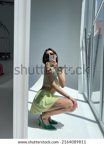 Fit tanned woman in light green beach skirt, sunglasses, crop top and heels, summer fashion stylish wear, take photo selfie on phone in mirror for social media, stories, vertical	