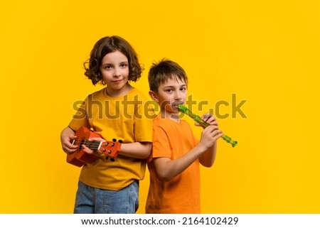 Musical duet in studio on yellow background. Child girl plays ukulele. School boy plays block flute. Two school children practice playing musical instruments Royalty-Free Stock Photo #2164102429