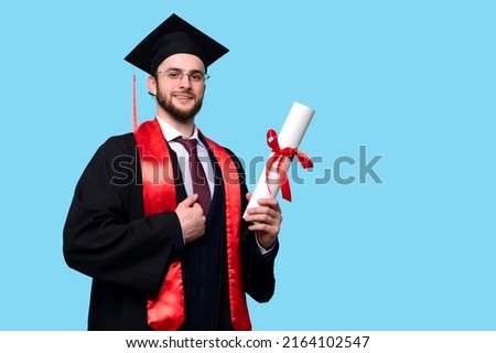 Graduate Male Student Wearing Ceremony Robe and Graduation Cap Holding Certificate on Blue Background in Studio. Man Celebrating Graduation and Getting Diploma.  Royalty-Free Stock Photo #2164102547