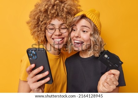 Two happy young women look gladfully at smartphone screen use banking application for paying online hold plastic credit card have toothy smile on face dressed casually isolated over yellow background