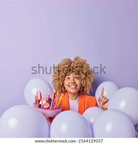Vertical shot of cheerful young woman with curly hair points index finger above shows something poses around inflated helium balloons holds delicious festive cake celebrates birthday anniversary