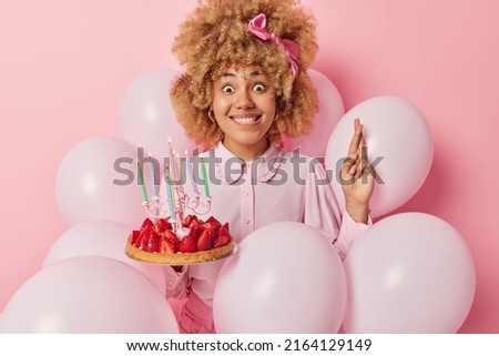 People holidays and celebration concept. Cute curly haired woman dressed in festive clothes holds delicious strawberry cake with burning candles keeps fingers crossed poses around inflated balloons