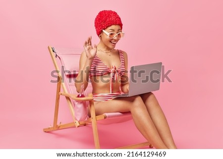 Positive woman waves hand and talks on video chat via laptop computer during livestream uses high speed internet for good connection dressed in swimwear sits on deck chait enjoys summer vacations