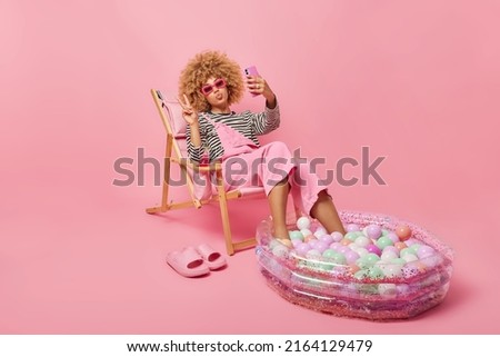 Lovely curly haired woman takes selfie makes peace gesture shares photos with friends in social networks enjoys summer time poses on deck chair keeps legs in inflated pool isolated on pink background Royalty-Free Stock Photo #2164129479