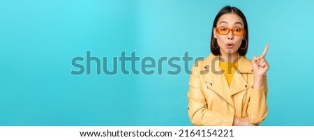 Excited asian girl raising finger, suggesting smth, eureka sign, has plan, standing in sunglasses over blue background