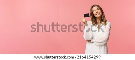 Portrait of cute tender feminine blond girl daydreaming about how she will waste her money on online shopping, look up dreamy, imaging as waiting delivery, hold credit card, pink background