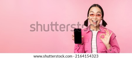 Enthusiastic young asian woman showing okay, ok sign, smiling pleased, mobile phone screen, smartphone application, standing over pink background