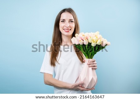 Smiling young woman holding vase with tulips on blue background.