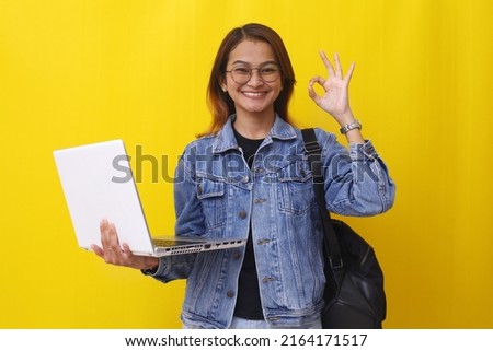 Cheerful asian college student standing while holding a laptop with okay hand sign gesture