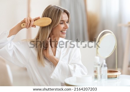 Smiling beautiful middle aged blonde woman in white silky bathrobe sitting in front of mirror at bathroom, combing her smooth hair with wooden brush at home, copy space