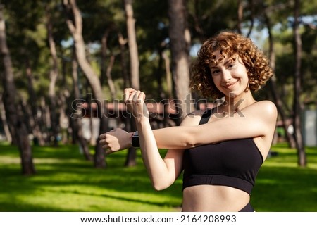 Beautiful redhead woman wearing black sports bra standing on city park, outdoors stretching her arms. Female doing warmup stretching workout while looking at the camera.